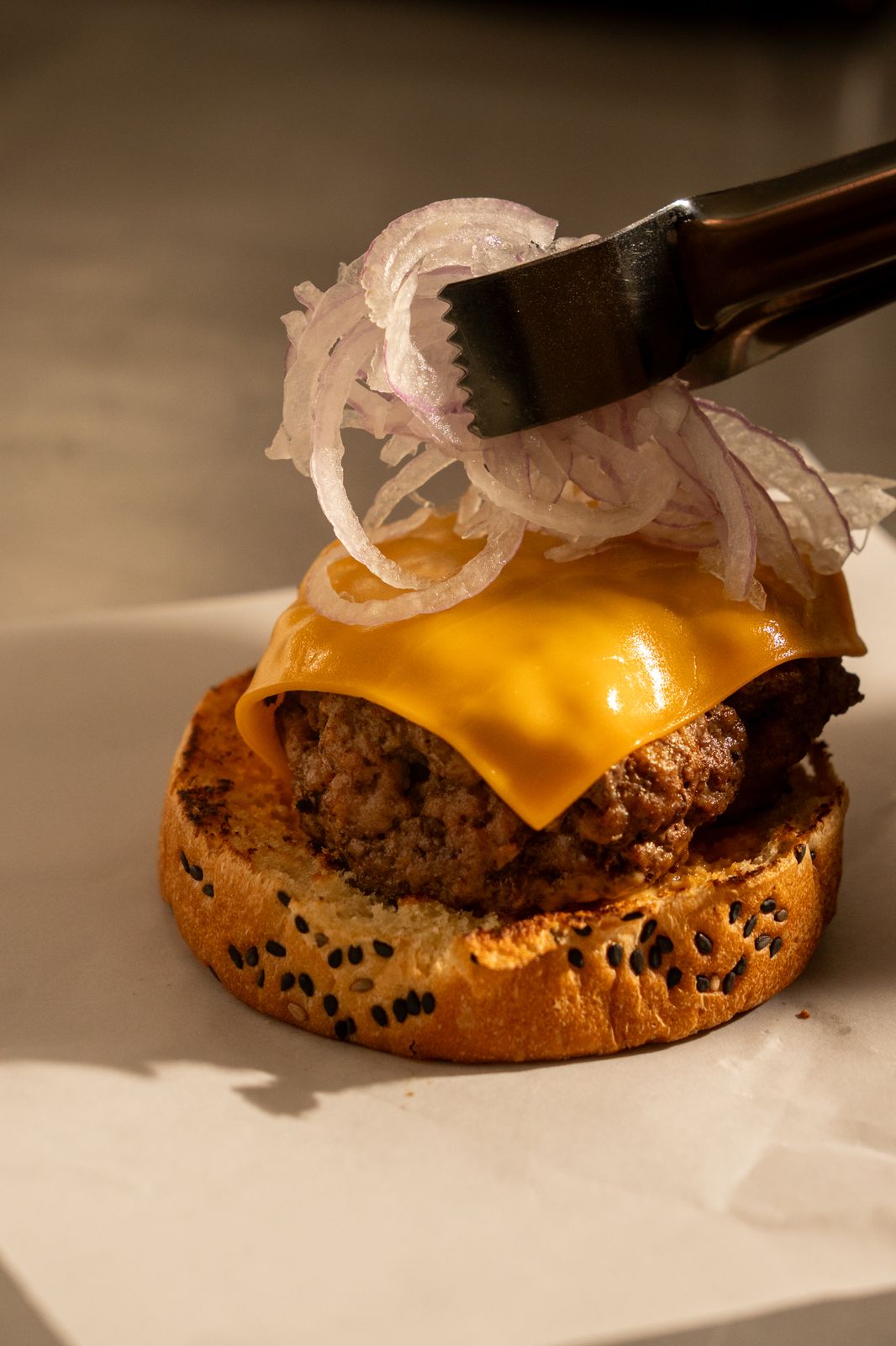 Tongs placing sliced onion on a cheeseburger with pickles